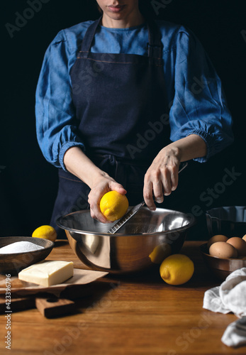 Woman in dark blue apron cooking lemon cake grating the lemon at the moment, front view