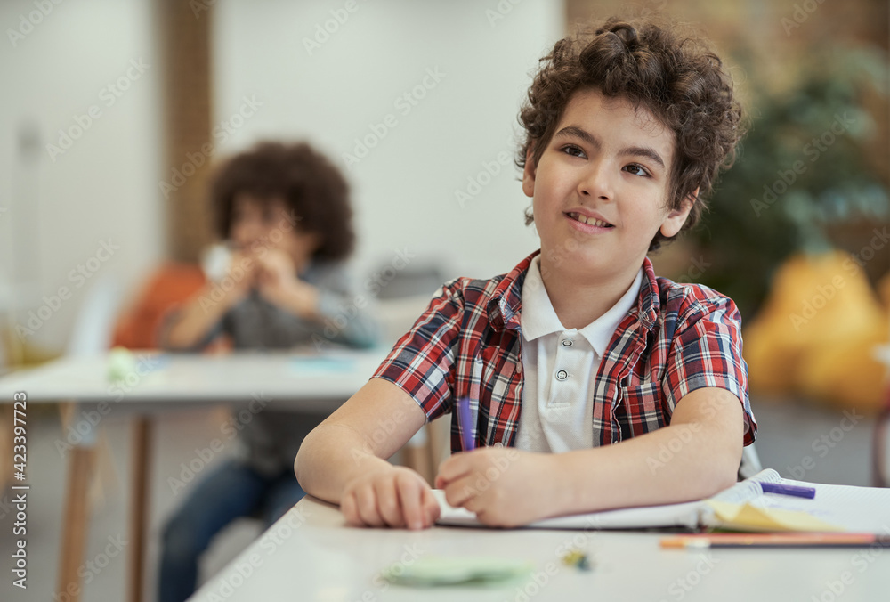 Naklejka premium Diligent little school boy listening to his teacher while sitting at the desk in classroom