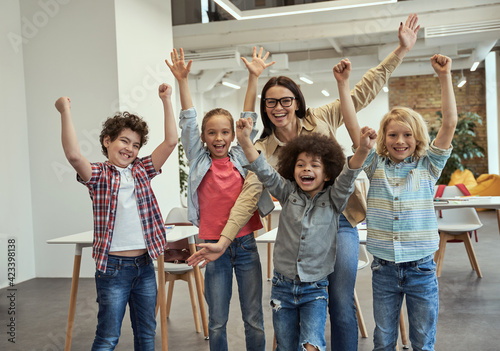 Studying is fun. Excited school team of four happy little pupils and their cheerful female teacher posing with arms raised together in the classroom