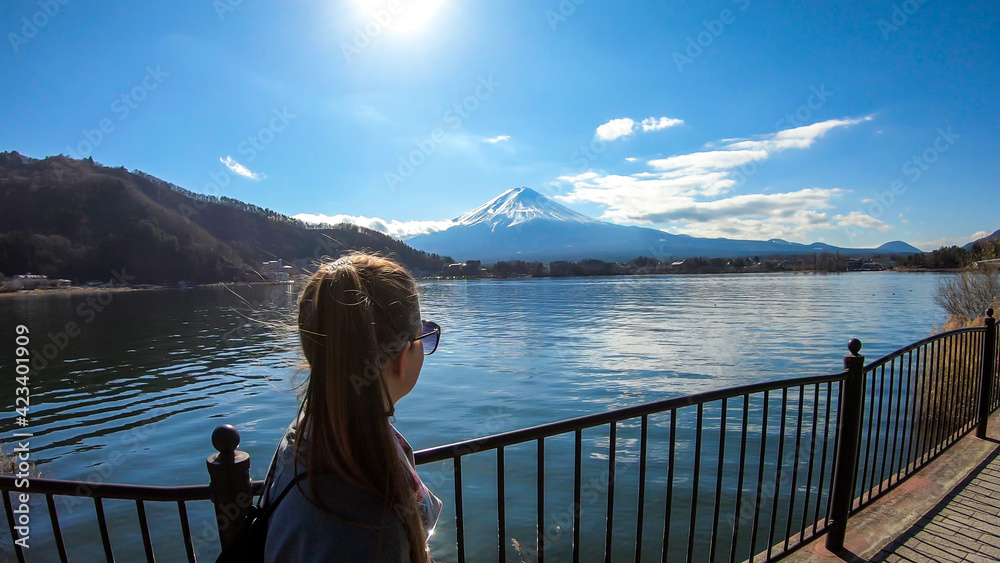 Woman walking across the bridge next to Kawaguchiko Lake, Japan with the view on Mt Fuji. The mountain is surrounded by clouds. Girl is enjoying the walk and good weather. Serenity and calmness
