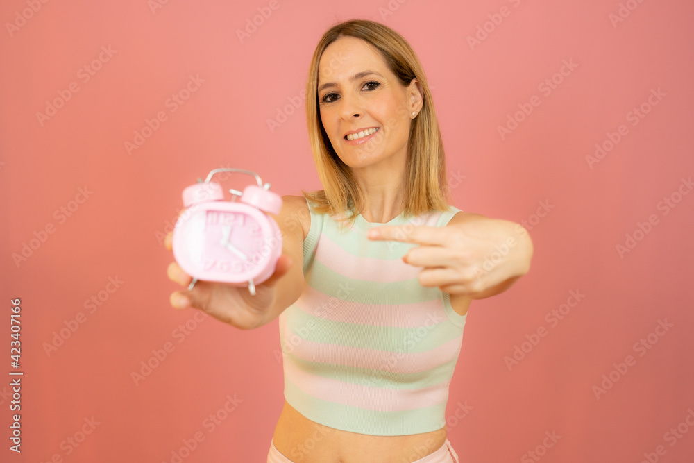 Young woman holding alarm clock standing over isolated pink background very happy pointing with hand and finger Young woman holding alarm clock standing over isolated pink background very happy pointing with hand and finger