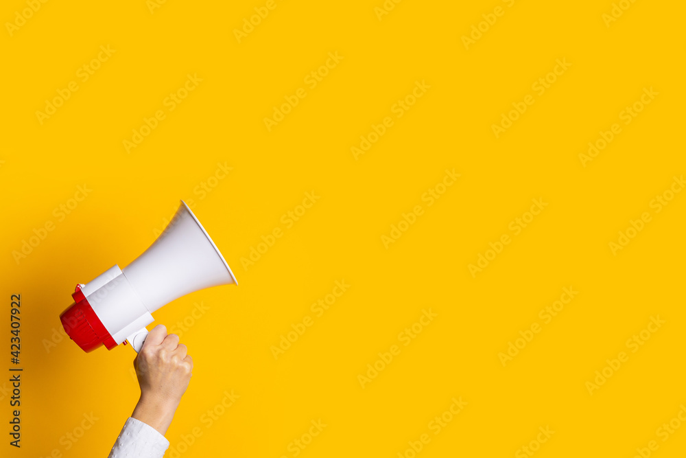 female hand holds a white with a red megaphone on a yellow background. - obrazy, fototapety, plakaty 