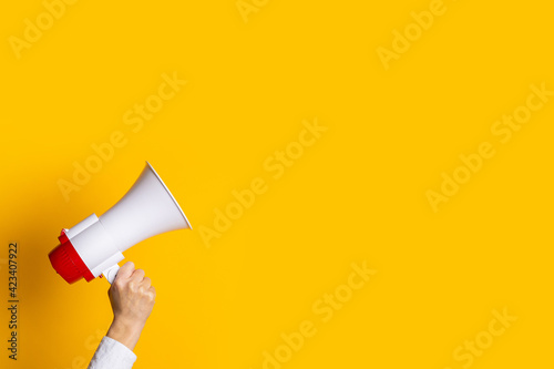 female hand holds a white with a red megaphone on a yellow background.