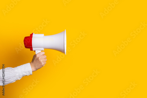 female hand holds a white with a red megaphone on a yellow background.