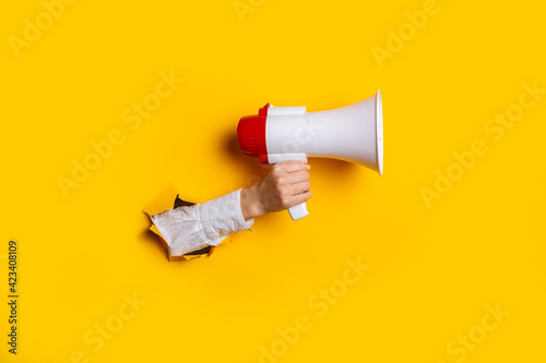 Hand holds a megaphone from a hole in the wall on a yellow background. Concept of hiring, advertising something. Banner photo