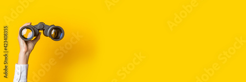 female hand holds black binoculars on a bright yellow background. Banner photo