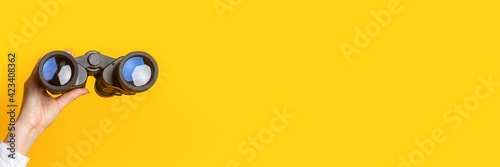 female hand holds black binoculars on a bright yellow background. Banner
