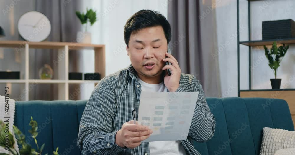 Likable confident in good mood adult chinese man in casual clothes sitting on the couch at home talking on smartphone with coworker simultaneously revisioning report Likable confident in good mood adult chinese man in casual clothes sitting on the couch at home talking on smartphone with coworker simultaneously revisioning report