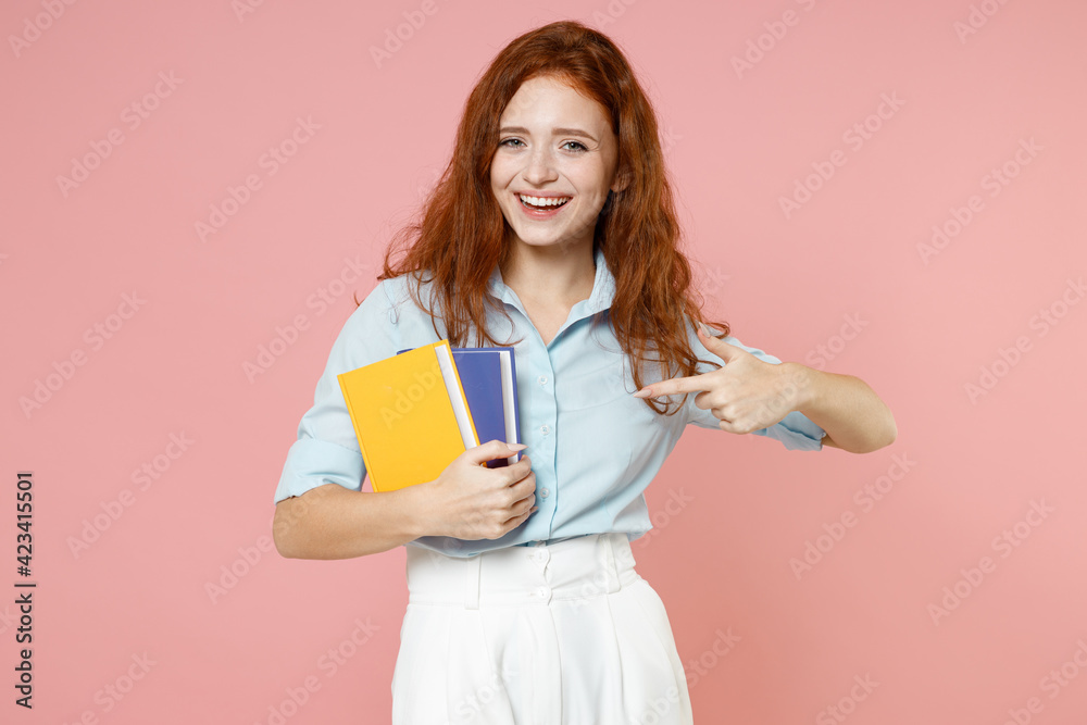 Young smiling smart redhead student woman in blue shirt hold books hold index finger on notebooks isolated on pastel pink background studio portrait Education high school university college concept. Young smiling smart redhead student woman in blue shirt hold books hold index finger on notebooks isolated on pastel pink background studio portrait Education high school university college concept.