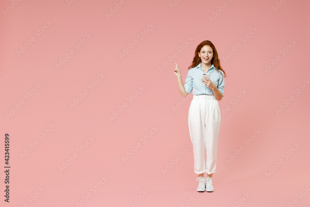 Full length side view of young smiling redhead woman in blue shirt pants using mobile cell phone point index finger aside on workspace area mock up isolated on pastel pink background studio portrait. Full length side view of young smiling redhead woman in blue shirt pants using mobile cell phone point index finger aside on workspace area mock up isolated on pastel pink background studio portrait.