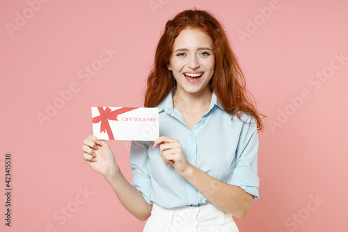 Young fun satisfied smiling happy caucasain ginger student redhead woman 20s wear blue shirt holding gift voucher flyer mock up looking camera isolated on pastel pink color background studio portrait.