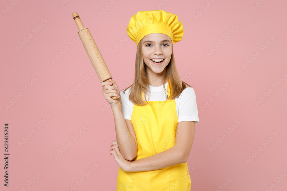 Teen happy girl chef cook confectioner baker housewife mother's helper in yellow apron white t-shirt toque cap hold rolling pin isolated on pastel pink background studio portrait. Food cake concept. Teen happy girl chef cook confectioner baker housewife mother's helper in yellow apron white t-shirt toque cap hold rolling pin isolated on pastel pink background studio portrait. Food cake concept.