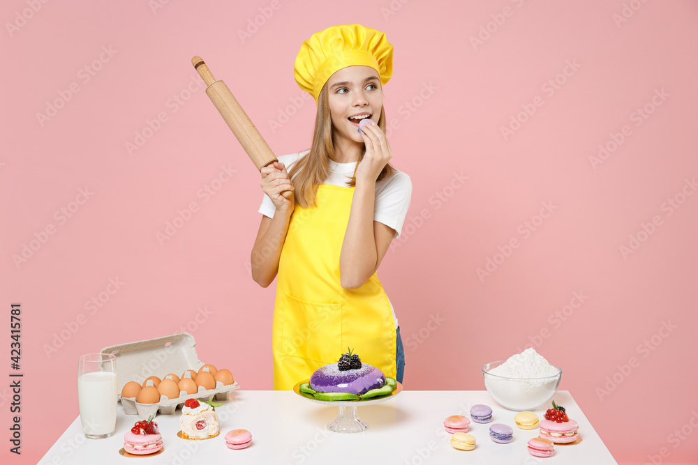 Teen happy girl chef cook confectioner baker wear yellow apron cap at table hold rolling pin eat macaroon isolated on pastel pink background studio Mousse cake cooking food process workshop concept. Teen happy girl chef cook confectioner baker wear yellow apron cap at table hold rolling pin eat macaroon isolated on pastel pink background studio Mousse cake cooking food process workshop concept.