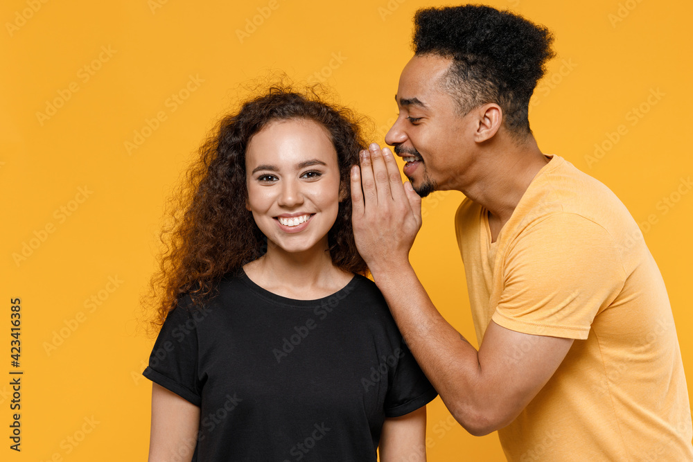 Young couple two friends family african surprised man woman in black t-shirt whisper gossip and tell secret behind her hand share news with girlfriend isolated on yellow background studio portrait. Young couple two friends family african surprised man woman in black t-shirt whisper gossip and tell secret behind her hand share news with girlfriend isolated on yellow background studio portrait.