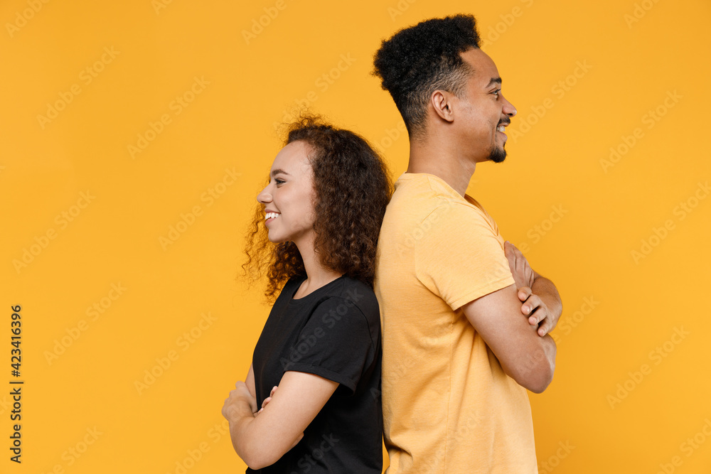 Side profile view young couple friends together team family african smiling happy man woman in black t-shirt stand back to back with hands crossed folded isolated on yellow background studio portrait. Side profile view young couple friends together team family african smiling happy man woman in black t-shirt stand back to back with hands crossed folded isolated on yellow background studio portrait.