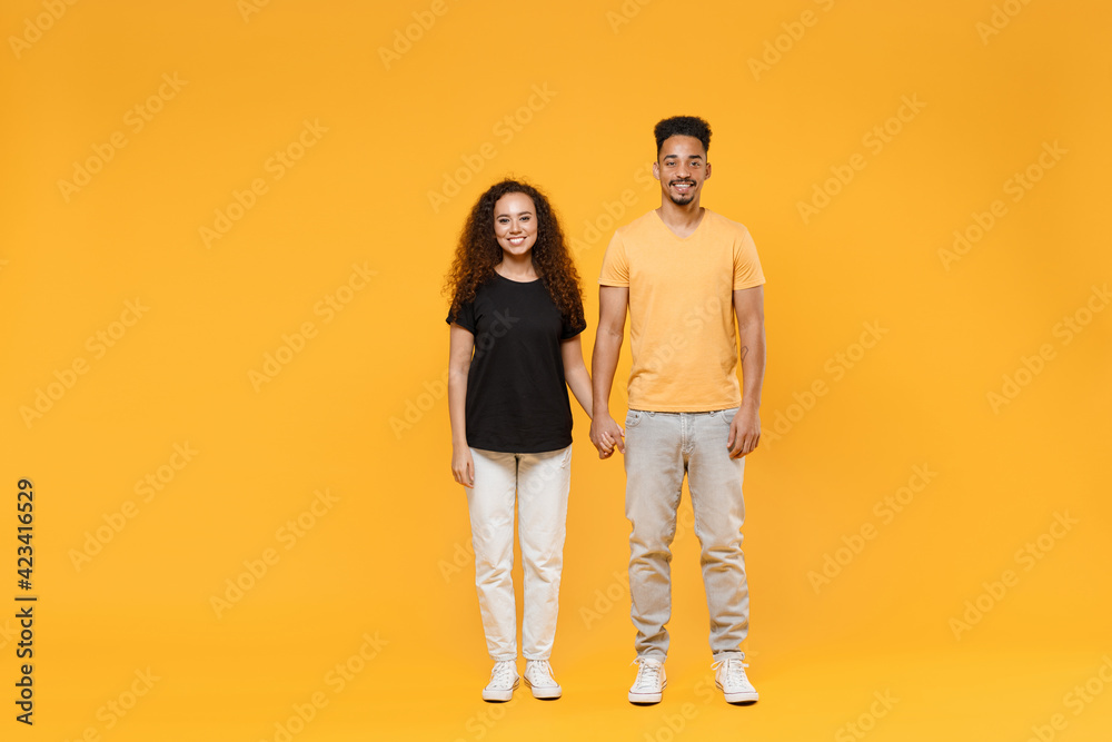 Full length young couple two fun american friends family cute happy fun smiling cheerful african man woman together in yellow black t-shirt hold hands isolated on orange background studio portrait Full length young couple two fun american friends family cute happy fun smiling cheerful african man woman together in yellow black t-shirt hold hands isolated on orange background studio portrait