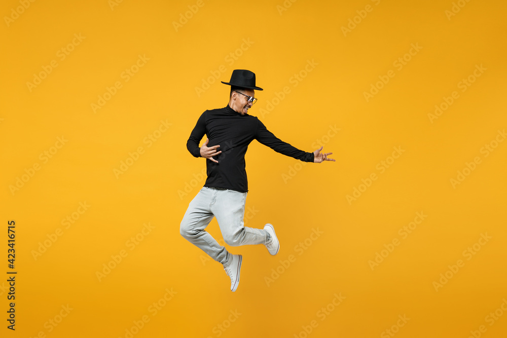 Full length of young active singer overjoyed fun happy excited fun african  man 20s in stylish black hat shirt eyeglasses jump high playing guitar  isolated on yellow color background studio portrait. Stock