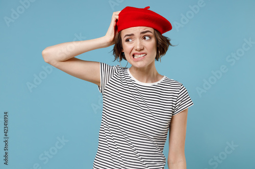 Young sad troubled pensive thoughtful woman 20s with short hairdo wearing french beret red hat striped t-shirt look aside sctratch head oops gesture isolated on pastel blue background studio portrait photo
