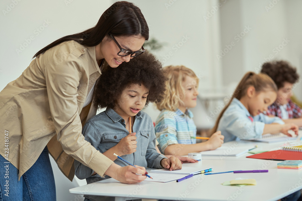 Helpful young female teacher showing how to do task to little schoolboy. Kids sitting at the table, studying in elementary school classroom