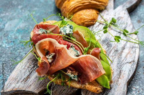 Croissant sandwich with prosciutto ham or jamon, blue cheese, avocado, microgin on blue background. Spanish breakfast, top view photo