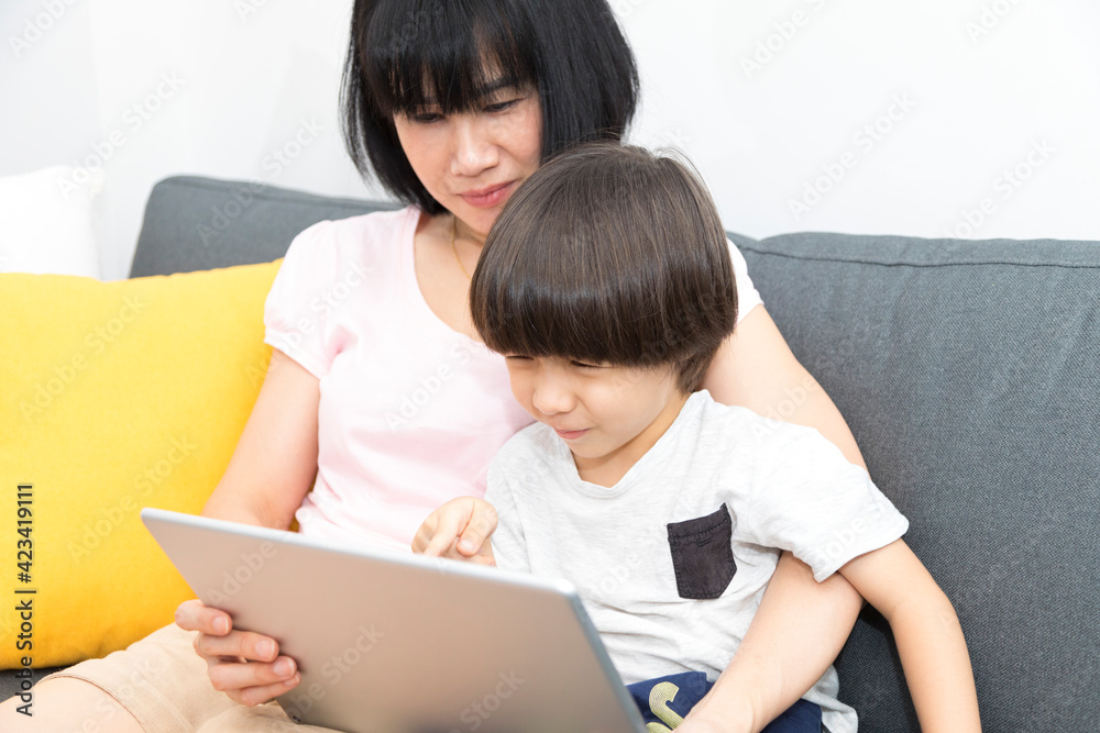 Cheerful Asian mother with little son sitting on sofa and playing video game on tablet while spending time together at home. Cheerful Asian mother with little son sitting on sofa and playing video game on tablet while spending time together at home.