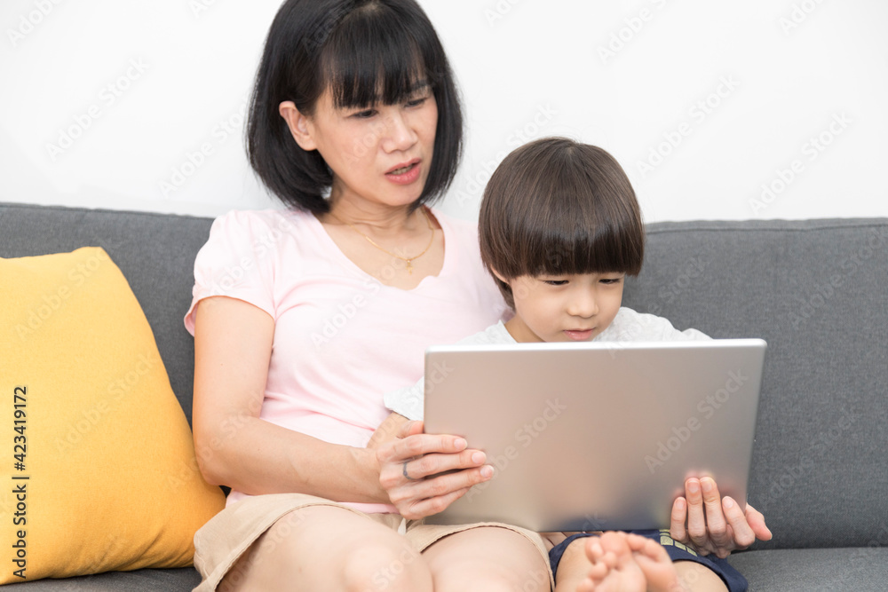 Cheerful Asian mother with little son sitting on sofa and playing video game on tablet while spending time together at home. Cheerful Asian mother with little son sitting on sofa and playing video game on tablet while spending time together at home.