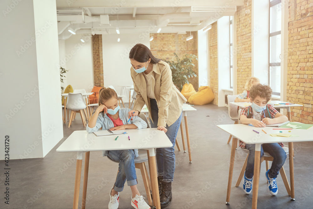 Full length shot of female teacher wearing protective face mask during coronavirus outbreak helping little girl in classroom. School kid studying in elementary school, sitting at the desk
