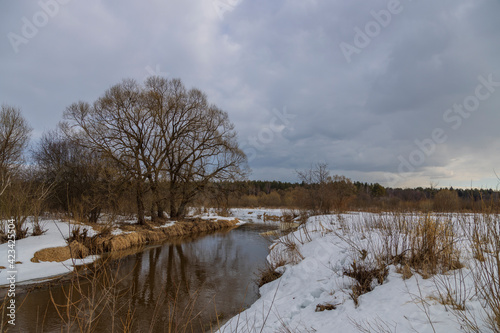 Spring landscape with a river and melting snow on the river bank. Dramatic sky. Large trees are reflected in the water. Early spring.