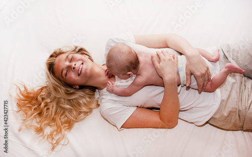 Mother and child on a white bed. Mom and baby boy or girl in diaper playing in sunny bedroom. Parent and little kid relaxing at home. Family having fun together. Bedding and textile for infant nursery