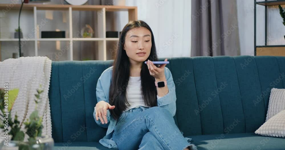Beautiful modern trendy young asian woman with black hair sitting on the soft couch in contemporary apartment and talking on smartphone on voice speaker Beautiful modern trendy young asian woman with black hair sitting on the soft couch in contemporary apartment and talking on smartphone on voice speaker
