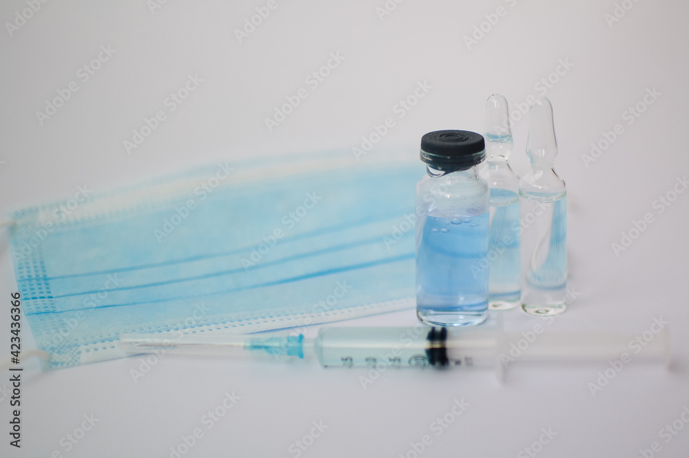 Vial vaccine, glass ampoules and a syringe with medical protective mask on white background, global vaccination concept Vial vaccine, glass ampoules and a syringe with medical protective mask on white background, global vaccination concept