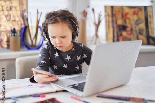A small kid girl in handsfree headphones sitting at a table using a laptop computer and smartphone. Doing business or small businesswoman concept. High quality photo