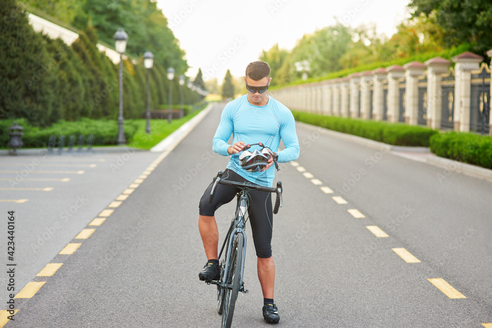 Fototapeta premium Full length shot of focused male cyclist wearing glasses, putting on helmet, getting ready for cycling, standing with his bike in the park on a daytime