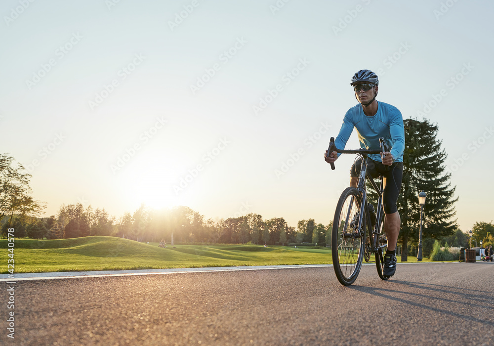 Full length shot of professional male cyclist in sportswear and protective helmet training, riding road bike in the park at sunset