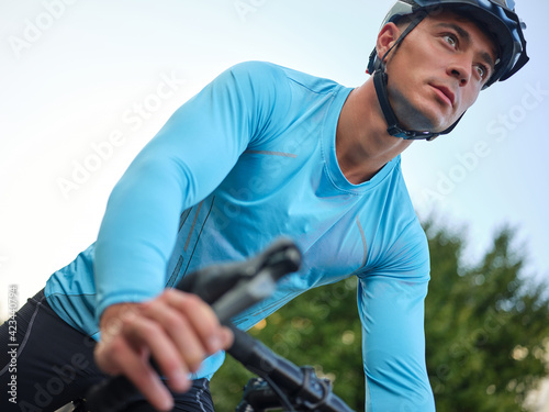 Low angle view of concentrated young male racer in sportswear and protective helmet looking away while riding his bike, cycling in park on a daytime