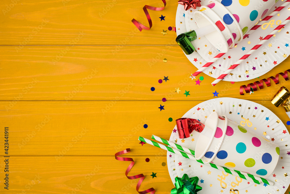 Overhead photo of cups drink with party tubules whistle multicolored confetti bow and plates isolated on the yellow wooden background with empty space Overhead photo of cups drink with party tubules whistle multicolored confetti bow and plates isolated on the yellow wooden background with empty space