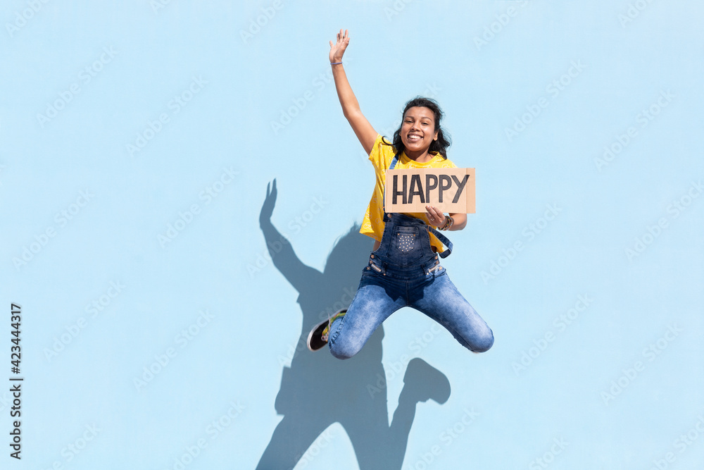 Smiling hispanic girl jumping with a banner with Happy text. She is isolated on blue background. Space for text. Smiling hispanic girl jumping with a banner with Happy text. She is isolated on blue background. Space for text.