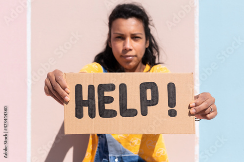 Close up of latin girl with serious gesture holding a banner with the text Help. She is next to a colorful wall. Selective focus.  © Alfredo López