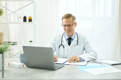 Cheerful doctor using laptop and filling in patient medical chart