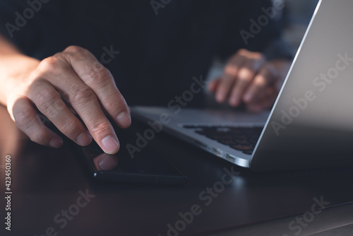Business man finger touching on mobile phone screen on table during working on laptop computer at office