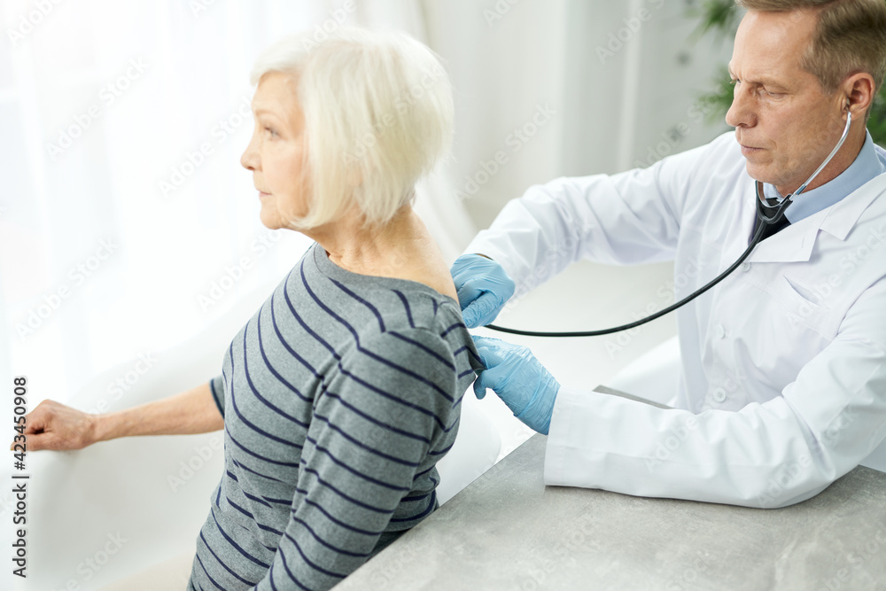 Obraz premium Serious doctor examining old woman with stethoscope in clinic