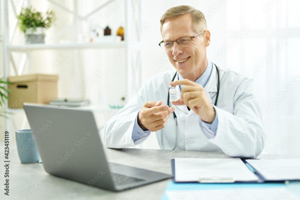 Cheerful doctor holding coronavirus vaccine and using laptop