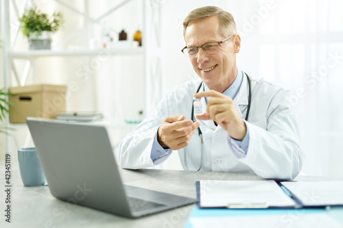 Cheerful doctor holding coronavirus vaccine and using laptop
