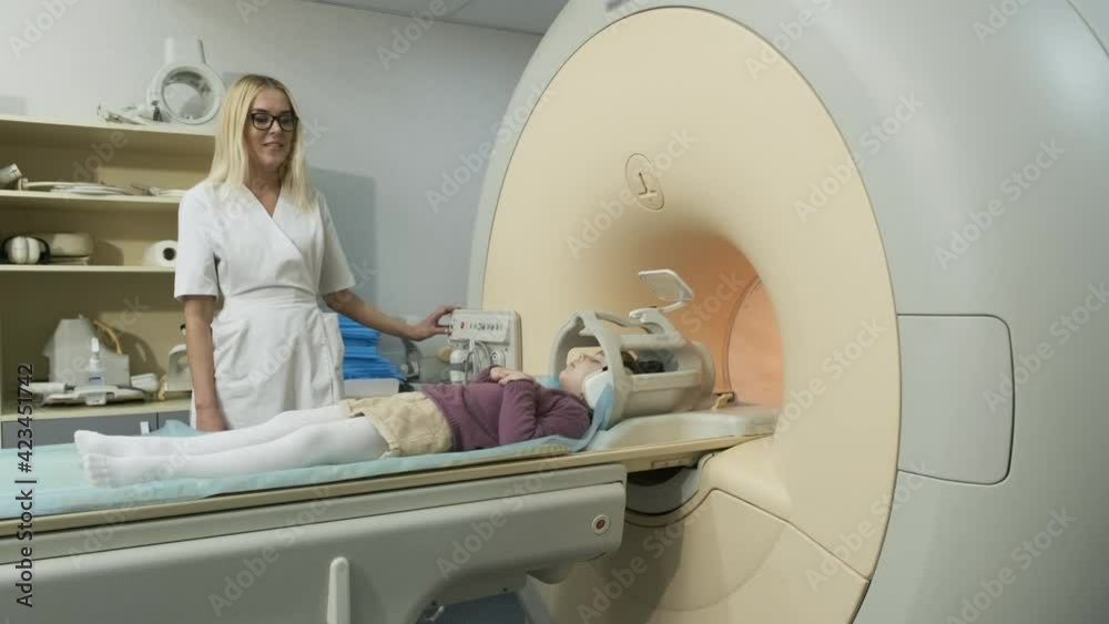 Woman doctor radiologist makes an MRI scanning of brain, head, neck for patient little girl, girl lies on automatic table, using modern equipment, coil on the head, headphones Woman doctor radiologist makes an MRI scanning of brain, head, neck for patient little girl, girl lies on automatic table, using modern equipment, coil on the head, headphones