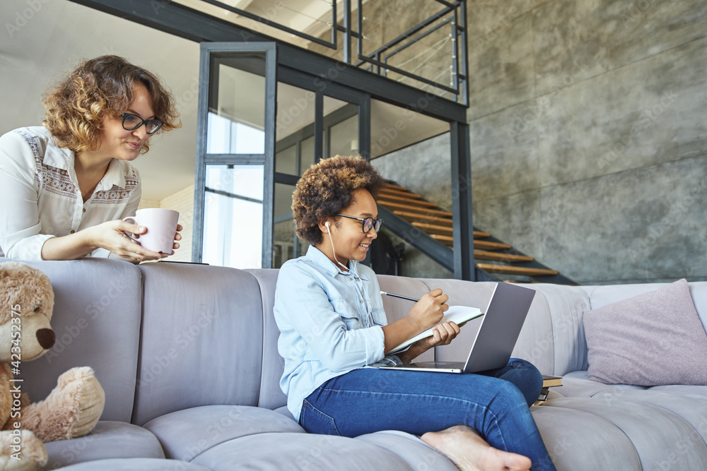 Adorable teen girl wearing earphones smiling, making notes, using laptop, sitting on the couch while spending time at home with mother during quarantine Adorable teen girl wearing earphones smiling, making notes, using laptop, sitting on the couch while spending time at home with mother during quarantine