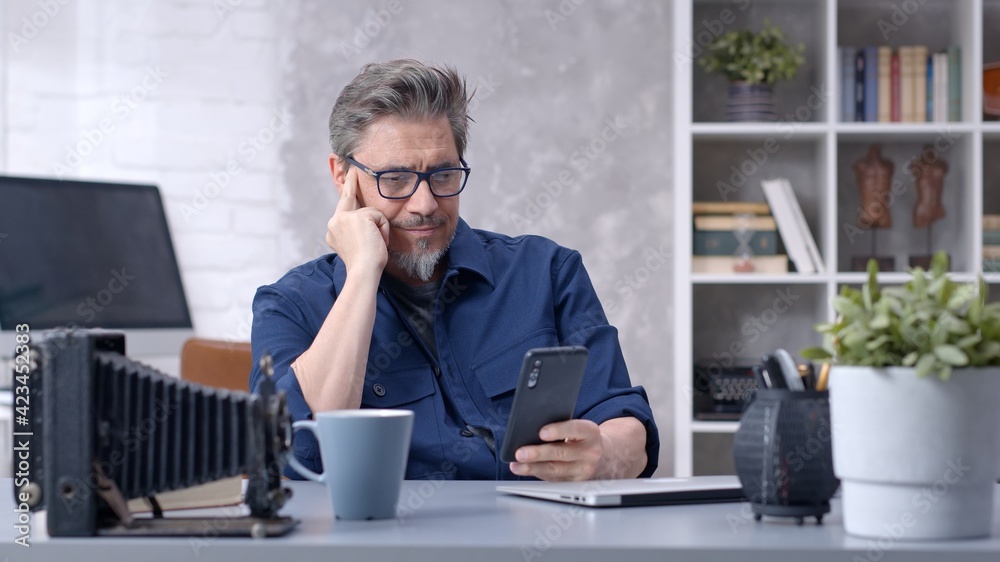 Bearded man working online with laptop computer and smart phone at home sitting at desk. Businessman in home office. Portrait of mature age, middle age, mid adult man in 50s. Bearded man working online with laptop computer and smart phone at home sitting at desk. Businessman in home office. Portrait of mature age, middle age, mid adult man in 50s.
