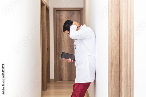 Latino doctor with a white coat and a stethoscope holding a tablet tired and leaning against a wall in a hospital corridor