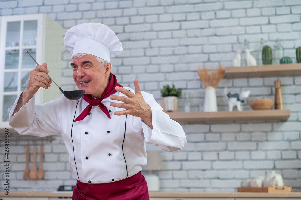 Portrait of smiling elderly chef tasting a scoop dish in the kitchen.
