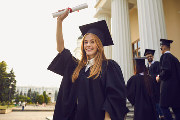 Happy smiling graduate girl dressed in black gown raises up her hand holding a diploma. Student posing in the university yard against the background of classmates. Success concept.