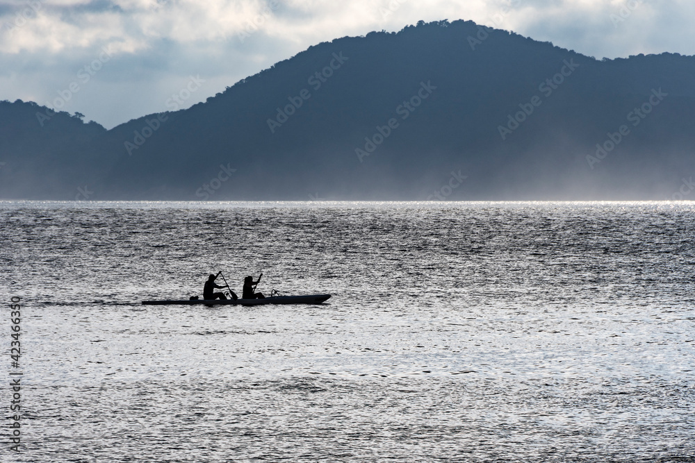 Fototapeta premium Two people canoeing in the late afternoon with a fog in the bay of the city of Santos. In the background, the mountains and blue sky.